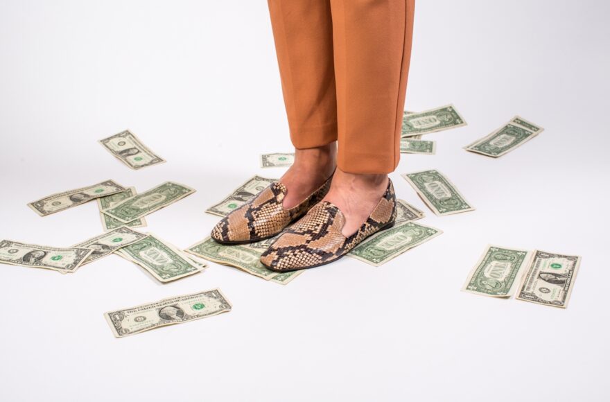 Person in rust-colored trousers and snakeskin loafers standing among scattered dollar bills, illustrating post-divorce financial planning.